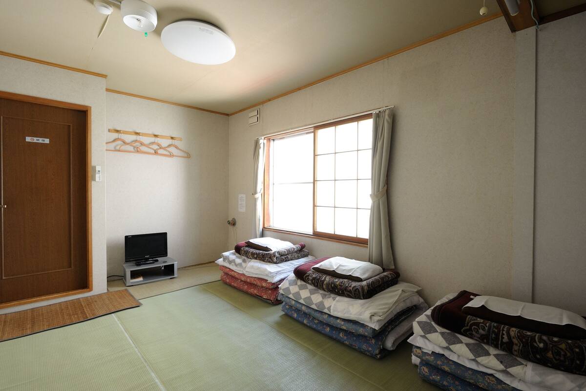 A room with tatami mat flooring, a television on a low stand, and a large window with curtains.