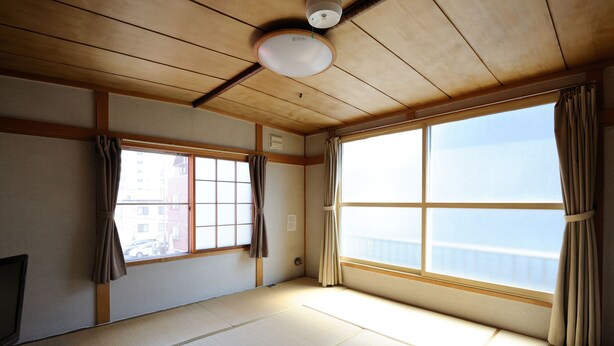 A traditional Japanese room with tatami flooring, large windows with curtains, and a ceiling fan. - Sapporo Inn NADA - Hostel (Sapporo)