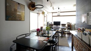A modern dining area with a long table, chairs, and a vase with flowers. There are ceiling lights and a large window in the background. - Sapporo Inn NADA - Hostel (Sapporo)
