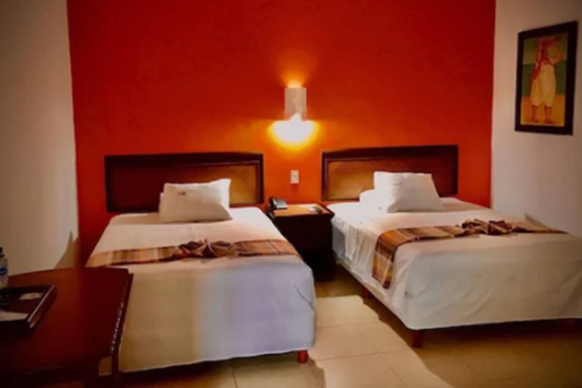 A hotel room with two beds, a nightstand, a desk, and a wall-mounted light.