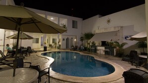 A swimming pool area with lounge chairs and umbrellas, surrounded by a building with a white exterior and a sun emblem. - Hotel María Jose (Mérida)