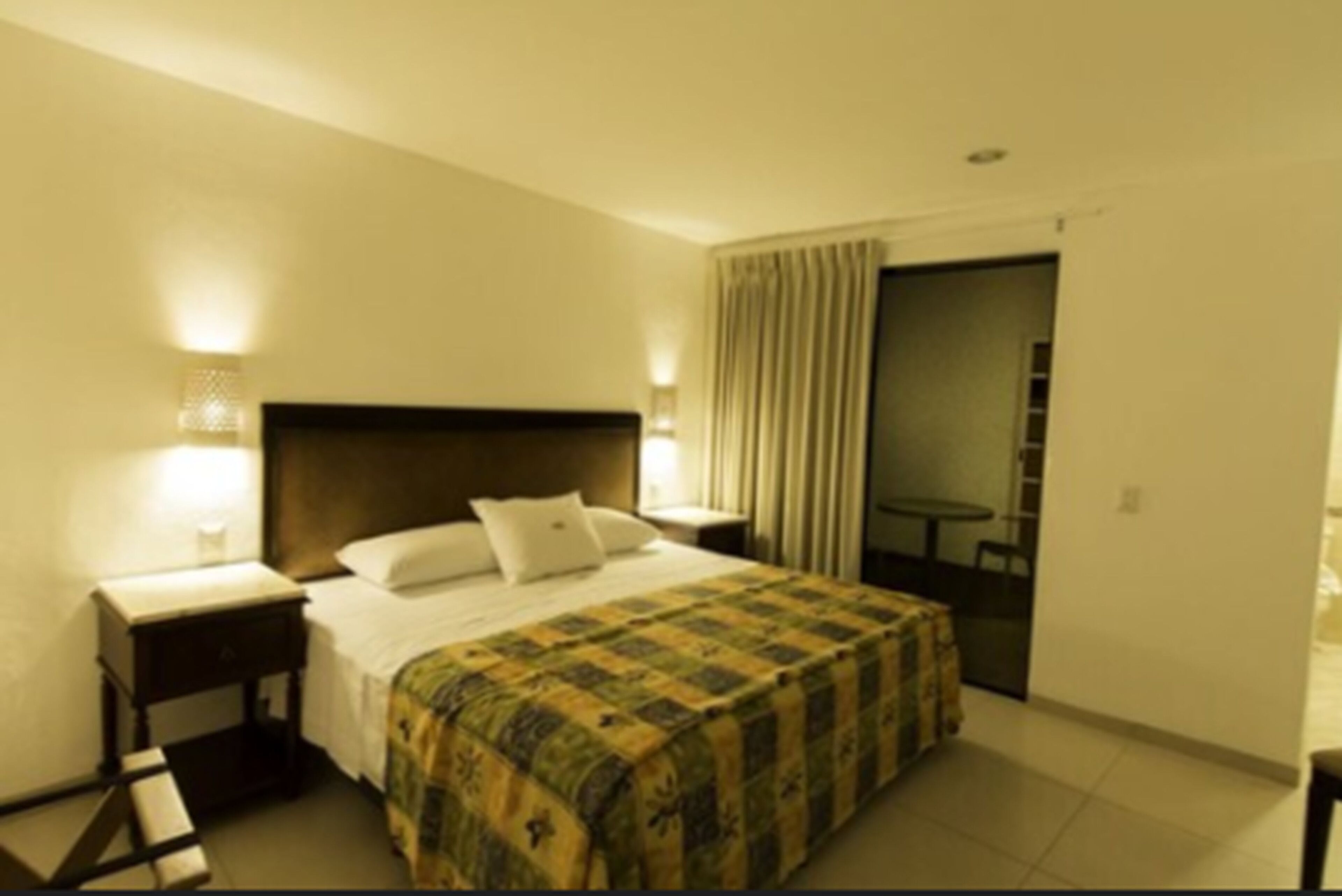 A hotel room with a bed, bedside tables, a small table, and a curtain.