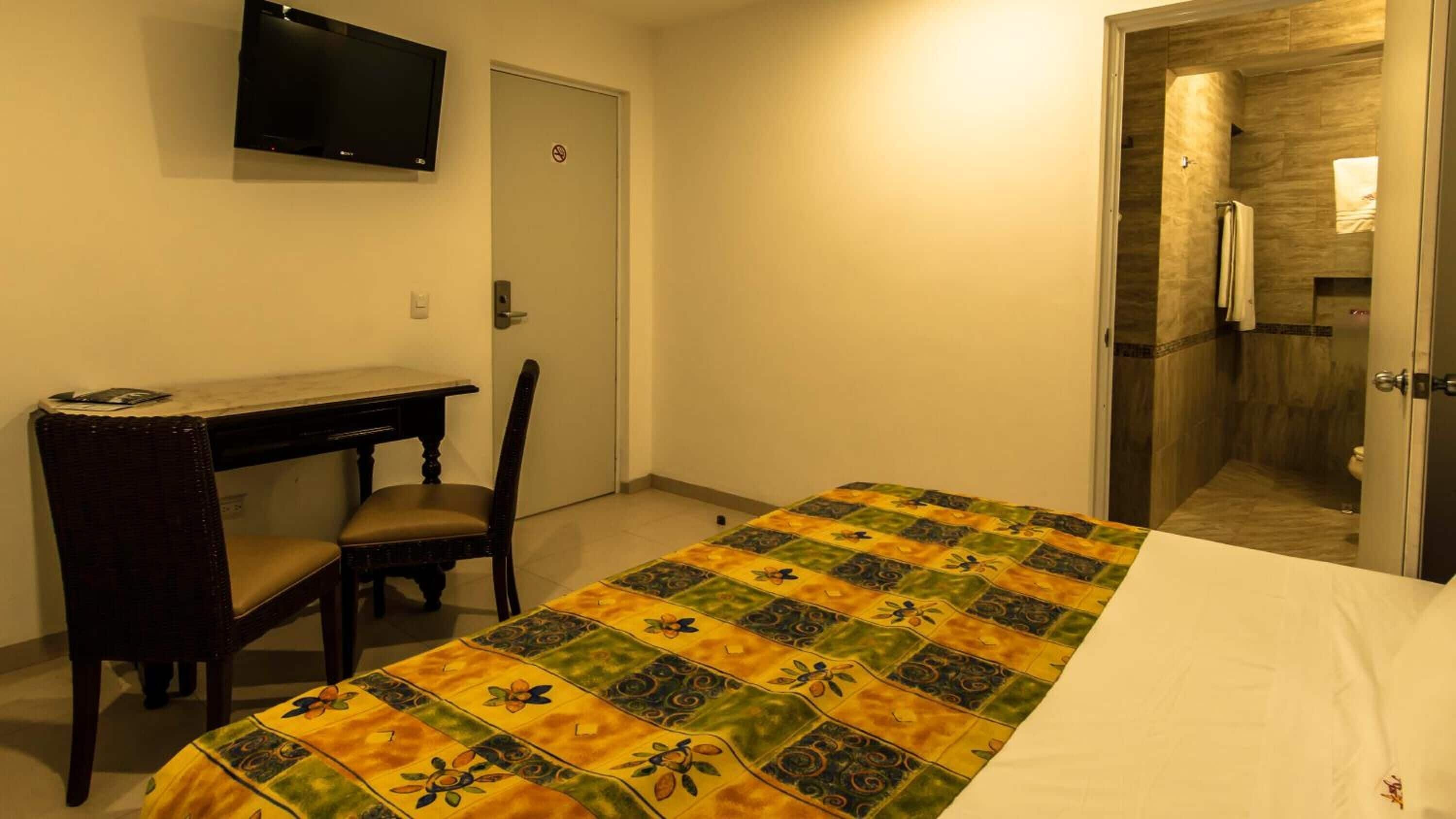 Habitación de hotel con cama, escritorio, silla, televisor y baño visible a través de una puerta abierta.