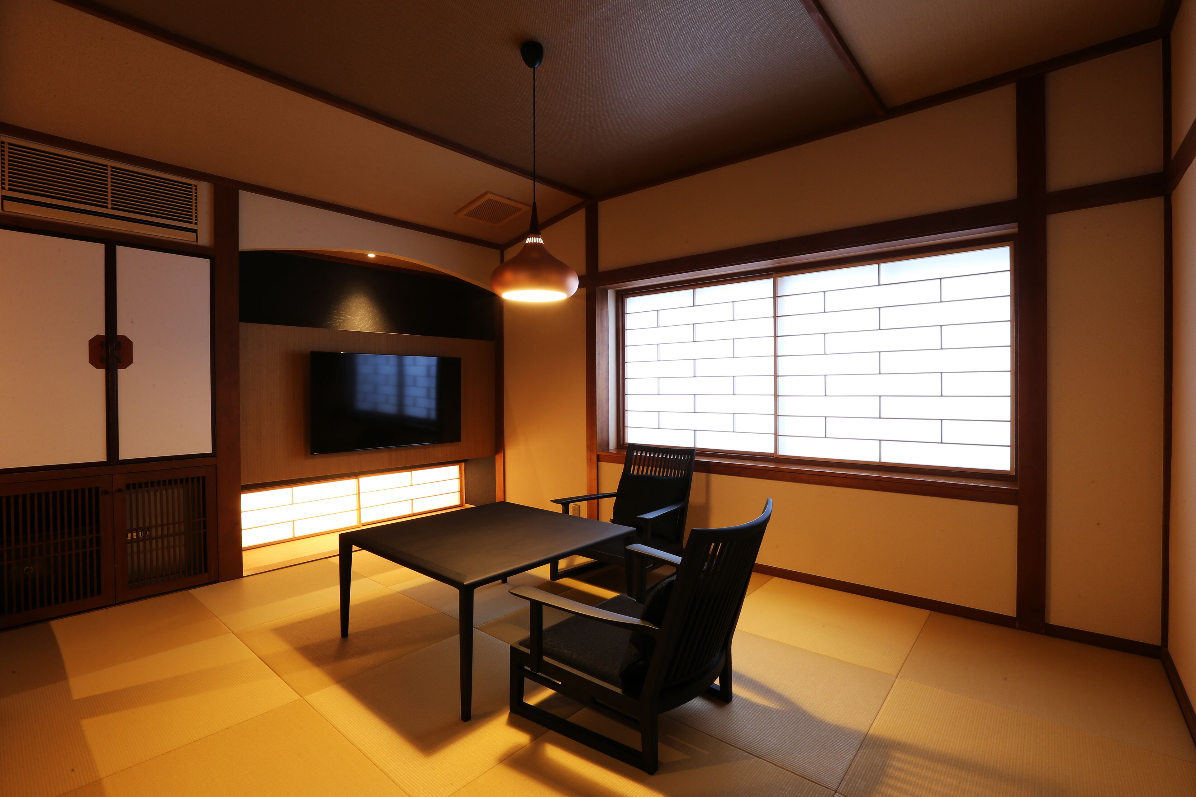 Une pièce de style japonais traditionnel, avec un sol recouvert de tatamis, une table basse, deux chaises, un téléviseur et une grande fenêtre donnant sur le ciel.
