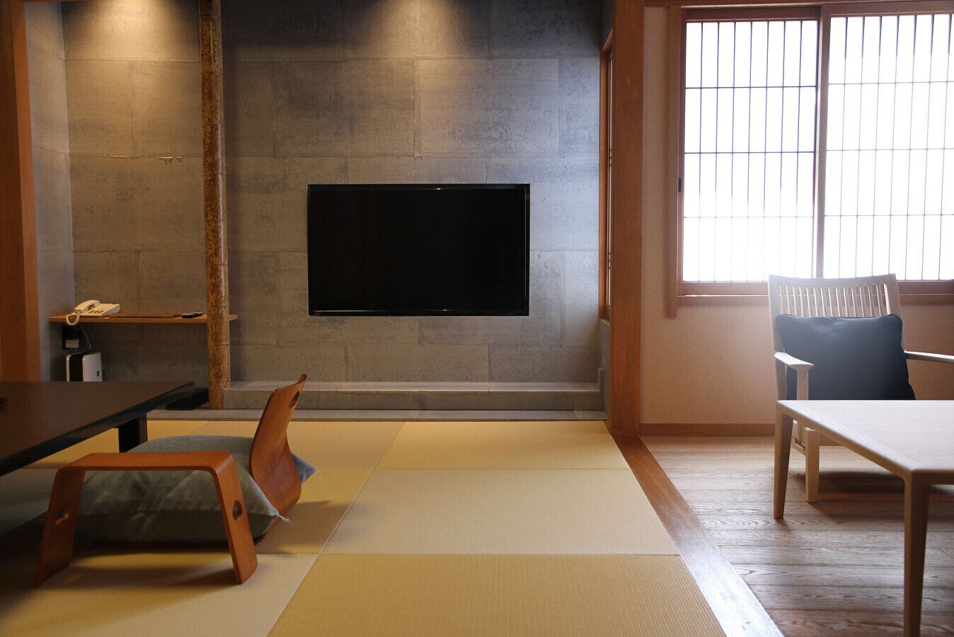 Een kamer met tatami matten, een aan een stenen muur bevestigde televisie, een houten tafel en een stoel met kussen.