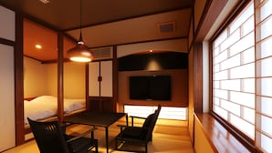 A traditional Japanese room with tatami flooring, a low table, two chairs, a bed, and a TV mounted on the wall. - Kinosaki Onsen Ichidaya (Toyooka)