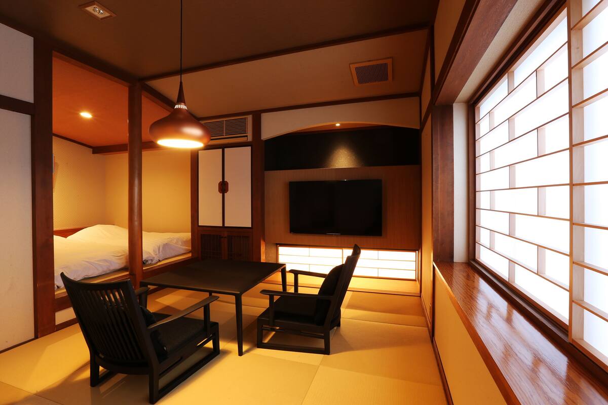 A traditional Japanese room with tatami flooring, a low table, two chairs, a bed, and a TV mounted on the wall.