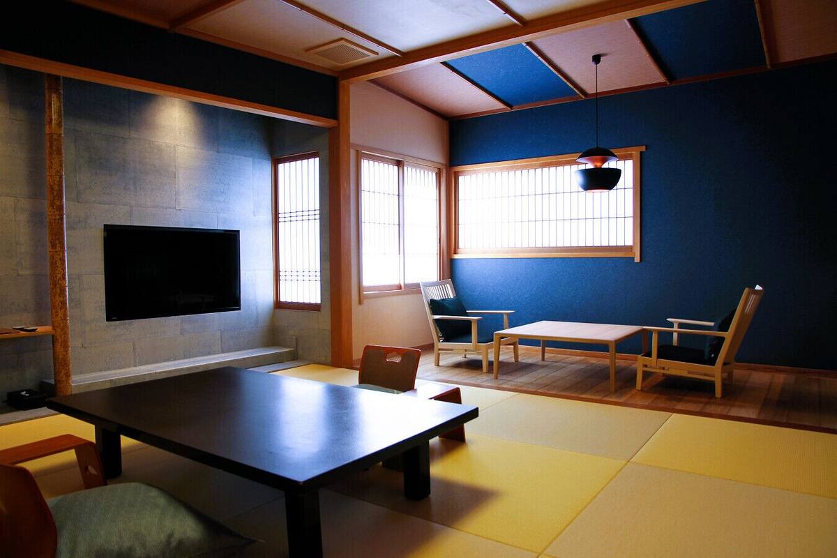 A traditional Japanese room with tatami flooring, a low table, a sofa, and a TV mounted on the wall.