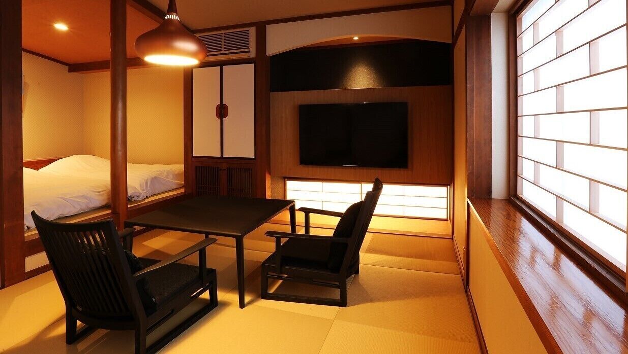 A traditional Japanese room with tatami flooring, a low table, two chairs, a bed, a television, and sliding doors.