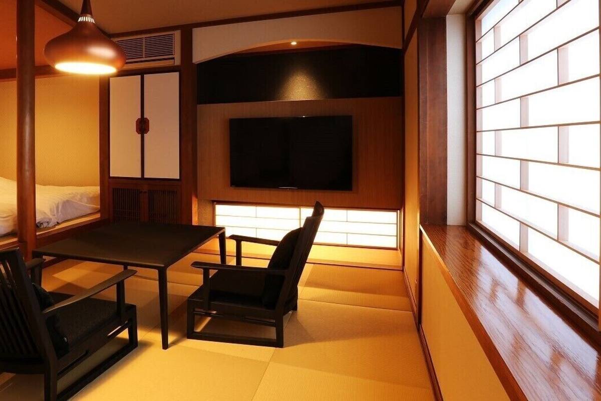 A traditional Japanese room with tatami flooring, a low table, two chairs, a bed, a television, and sliding doors.