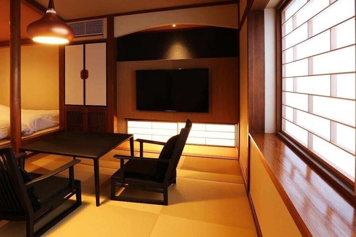 A traditional Japanese room with tatami flooring, a low table, two chairs, a bed, a television, and sliding doors.