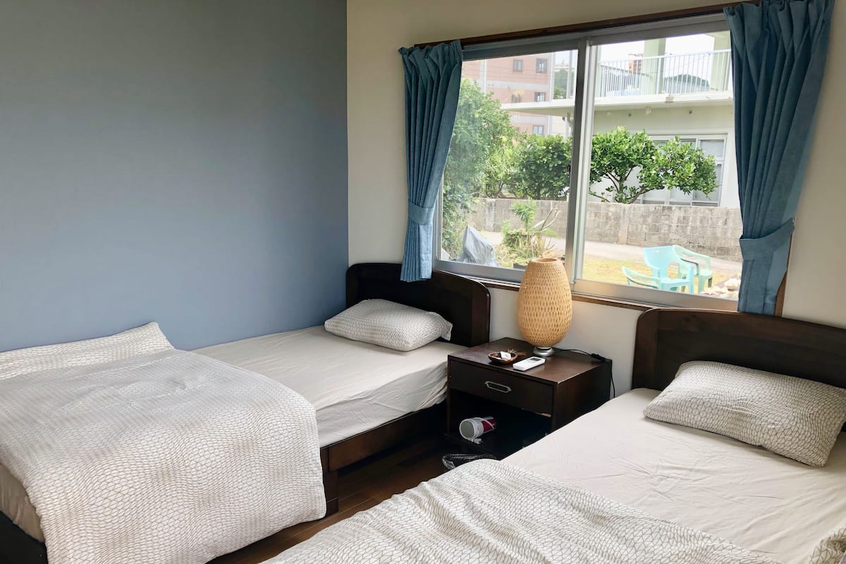 Quarto de hotel com duas camas, mesa de cabeceira com abajur e janela com cortinas.