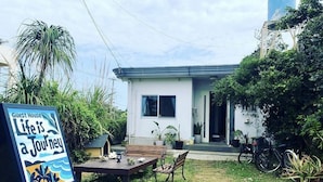 A guest house with a patio area featuring a table and chairs, surrounded by potted plants and a bicycle. - Life is a Journey - Hostel (Yomitan)