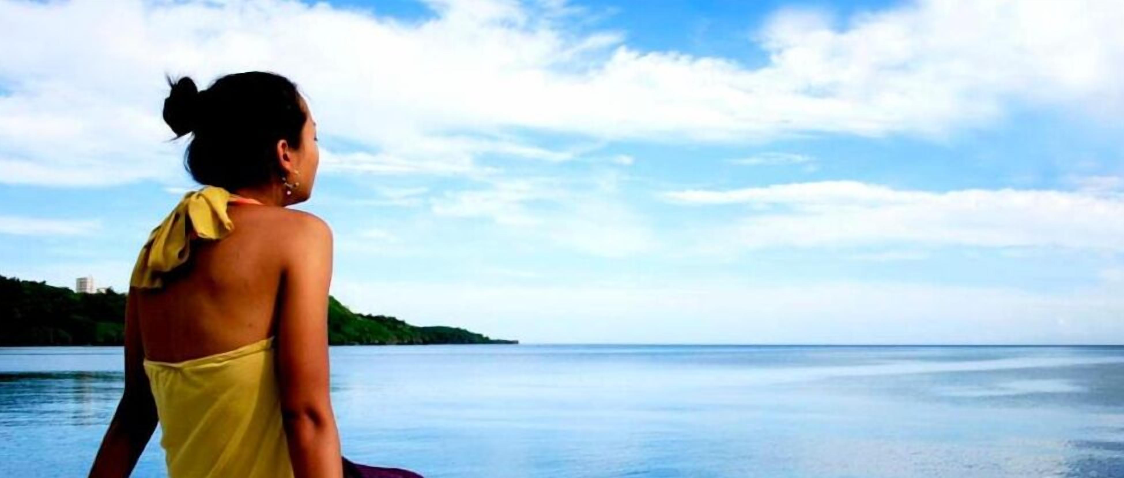 A person in a yellow garment overlooking a calm body of water with a distant landmass and a clear sky.