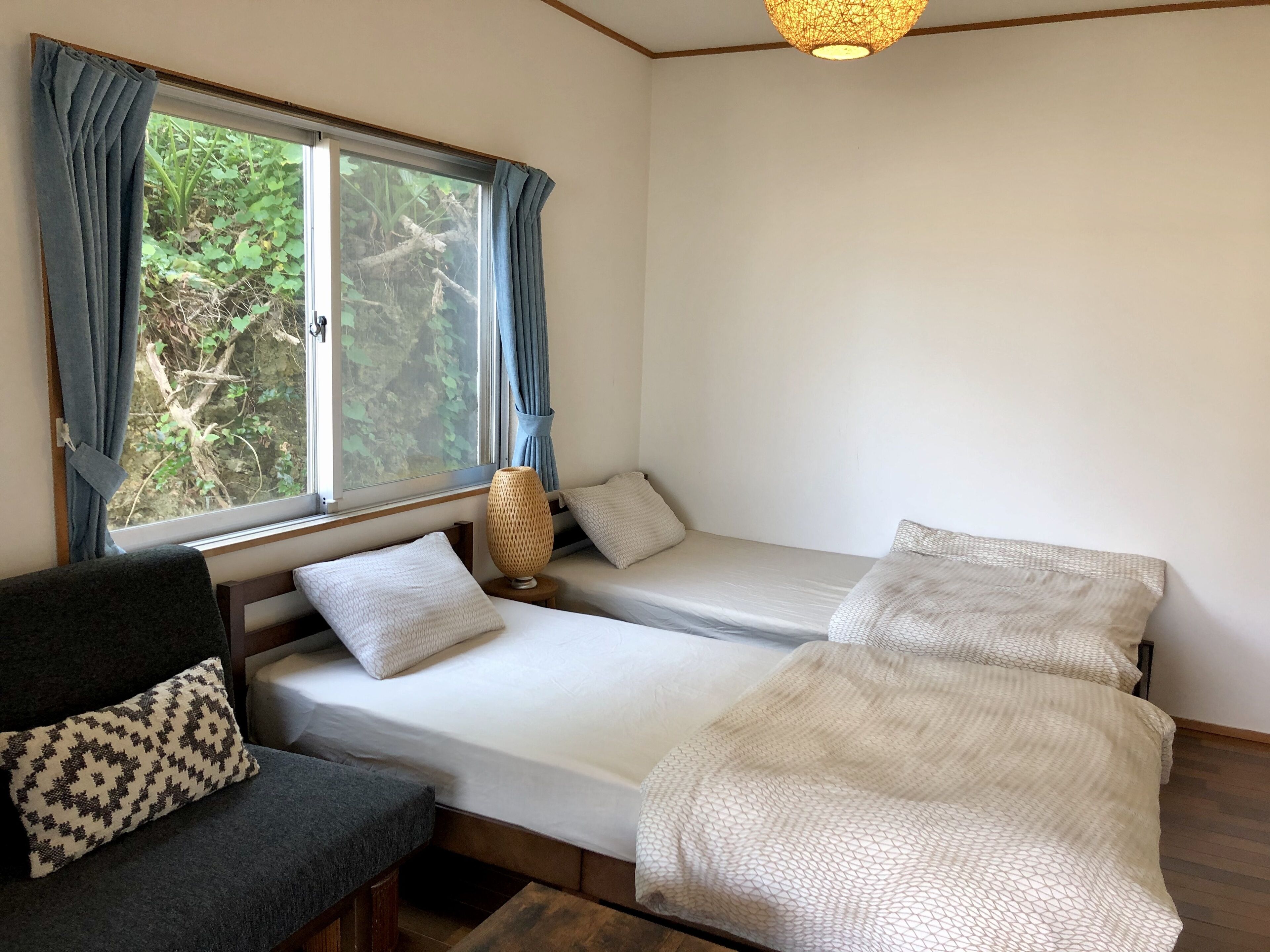 Quarto com duas camas de solteiro, um sofá, janela com cortinas e luminária pendente.