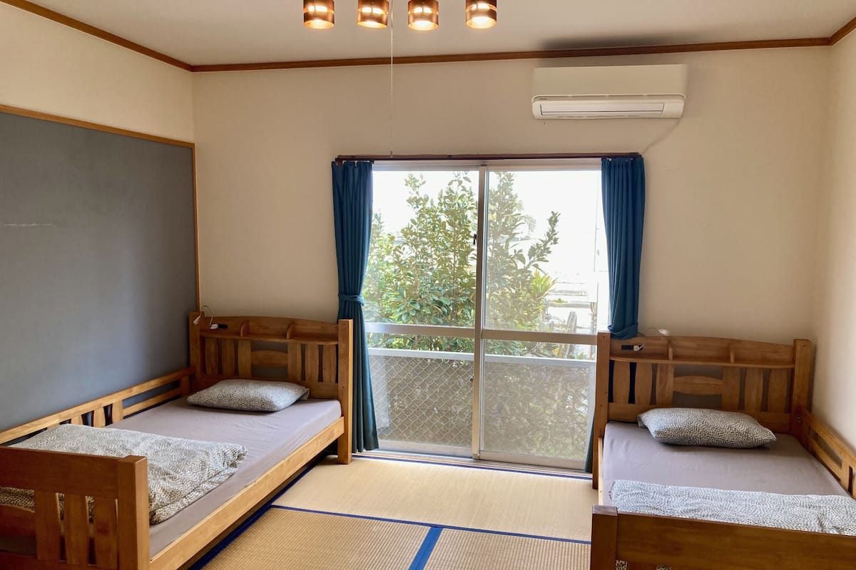 Quarto com duas camas de madeira, uma janela grande com cortinas azuis, aparelho de ar condicionado e piso de esteira de tatame.