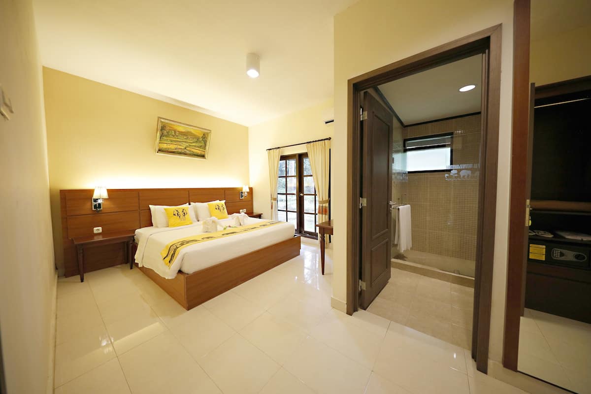 Une chambre d’hôtel avec un grand lit, des tables de chevet, une petite table, un tableau au mur et une salle de bain visible à travers une porte ouverte.