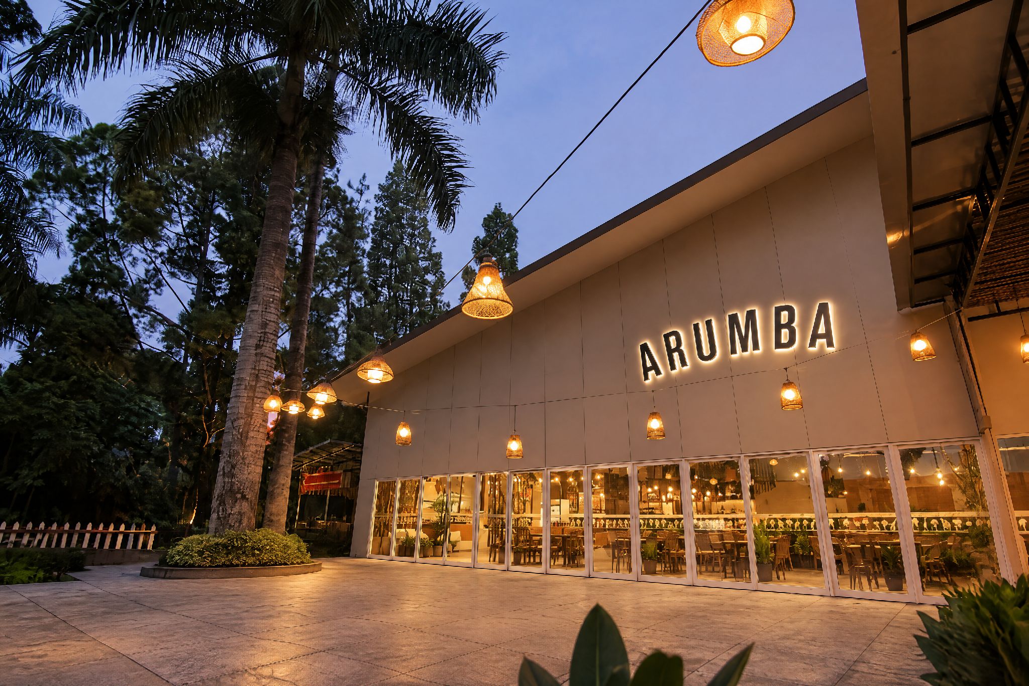 A building with the name 'ARUMBA' illuminated on the facade, surrounded by trees and outdoor seating.