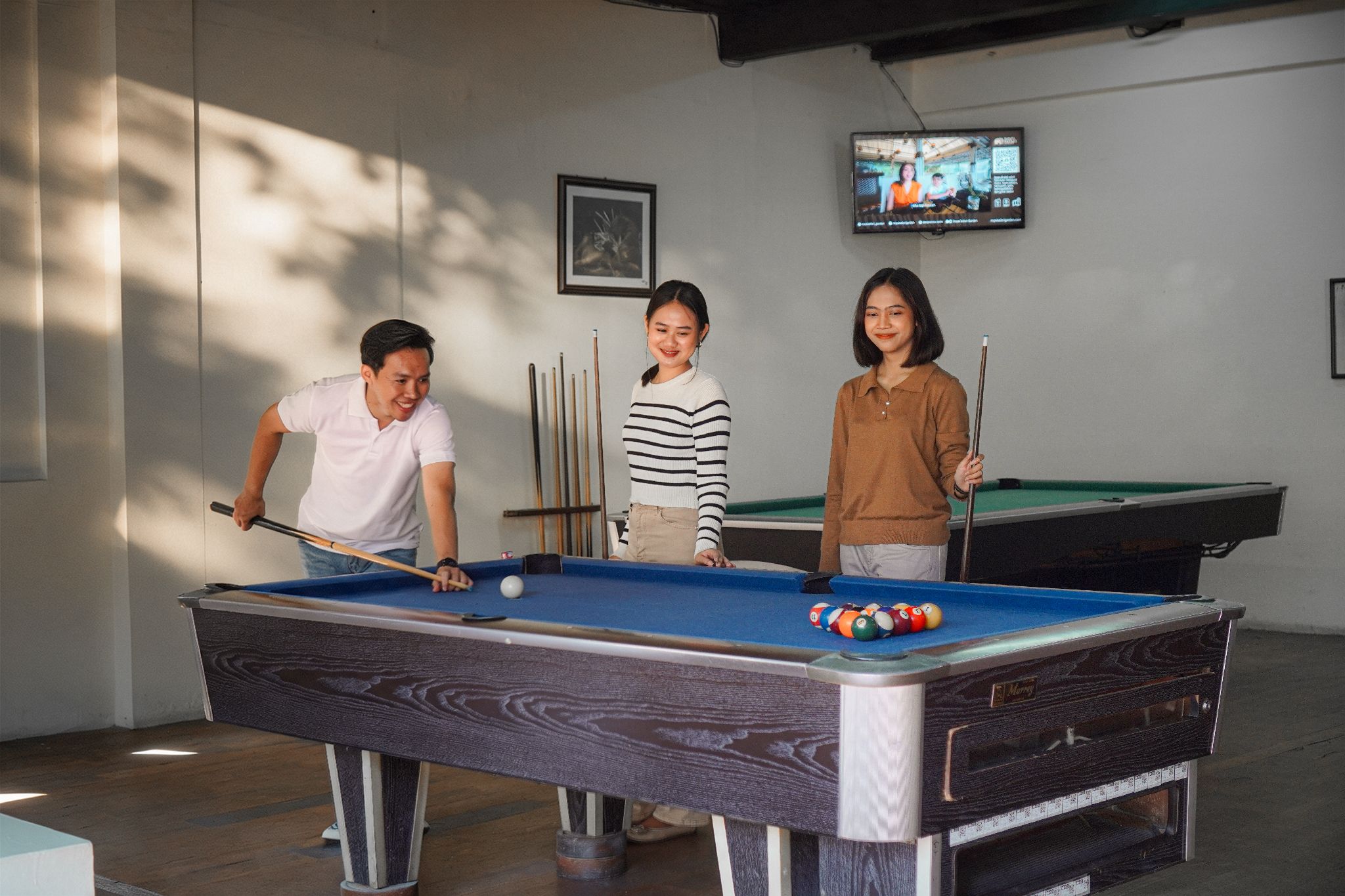 A pool table with three people playing, a television screen in the background, and a framed picture on the wall.