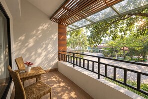 Un balcon donnant sur une rue et des arbres, une chaise en osier, une petite table avec une plante et une balustrade en bois.