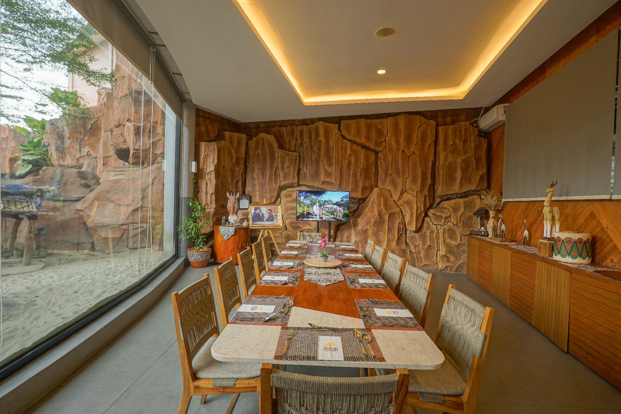 A dining area with a long table set for multiple people, surrounded by chairs, and a television mounted on the wall.