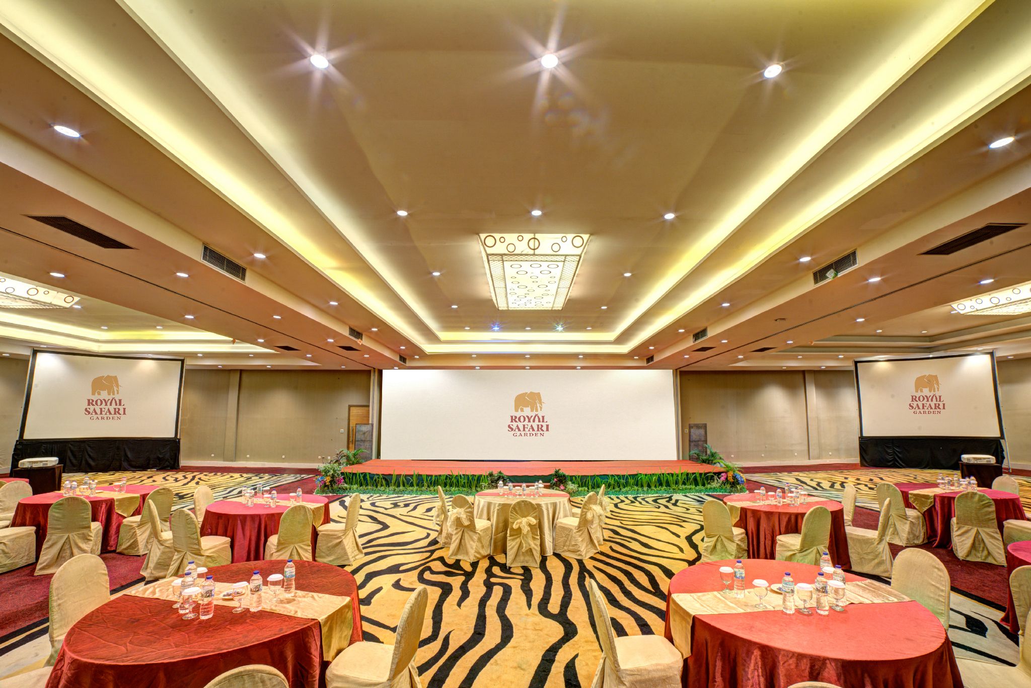 A banquet hall with round tables, chairs, and a stage with a presentation screen.