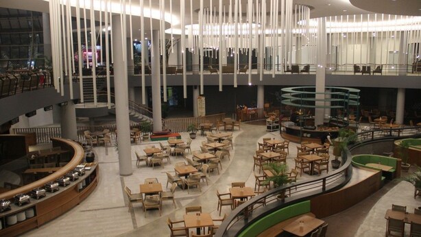 A spacious indoor area with multiple levels, featuring a central open space surrounded by tables and chairs, and a curved seating area with a green accent wall.