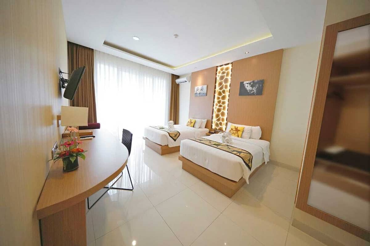 Une chambre d’hôtel avec deux lits, un bureau et une chaise. Il y a une fenêtre avec des rideaux, une lampe fixée au mur et une petite table sur laquelle est disposé un arrangement floral.