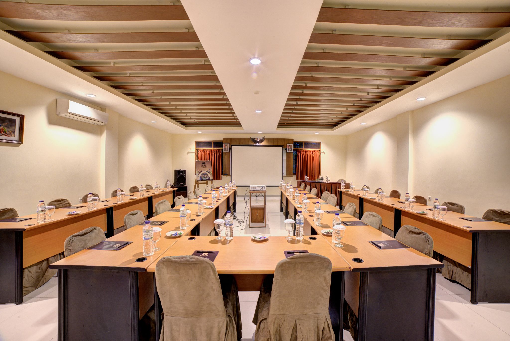A conference room with long tables, chairs, and a large screen at the front.