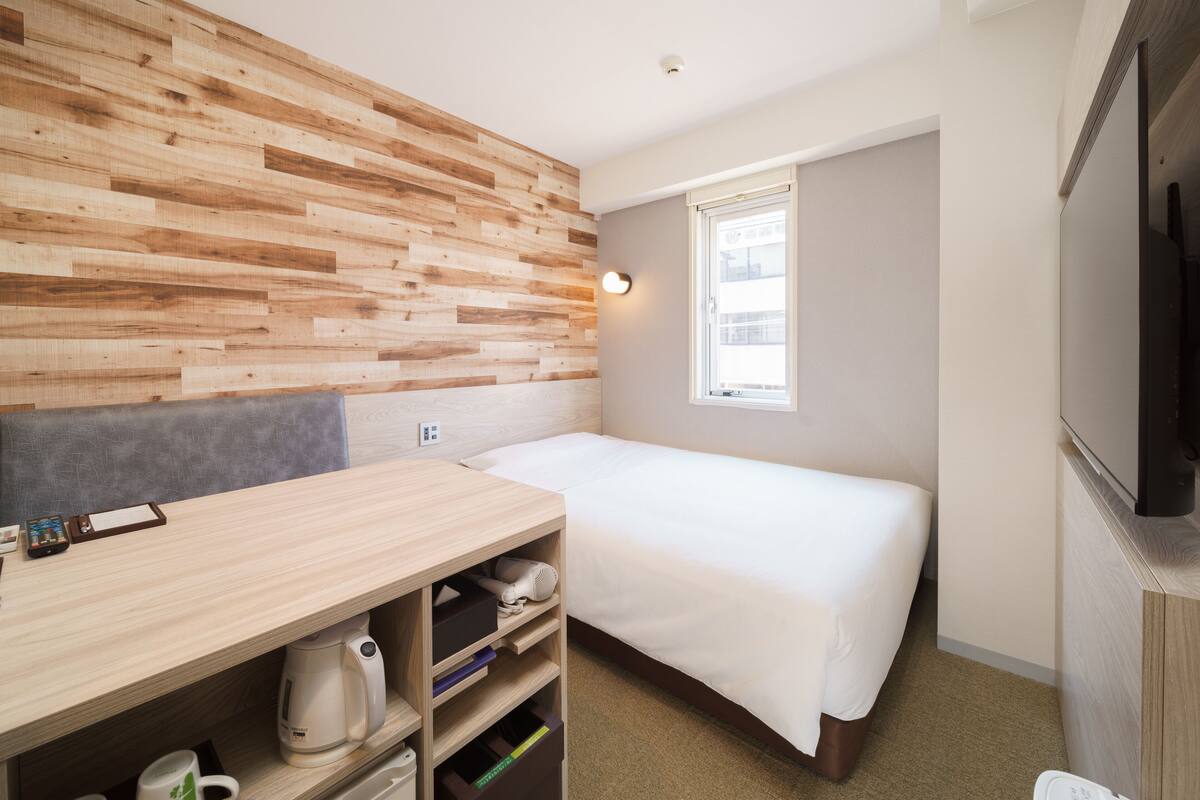 A modern hotel room with a wooden accent wall, a bed, a desk with a coffee maker, and a TV.