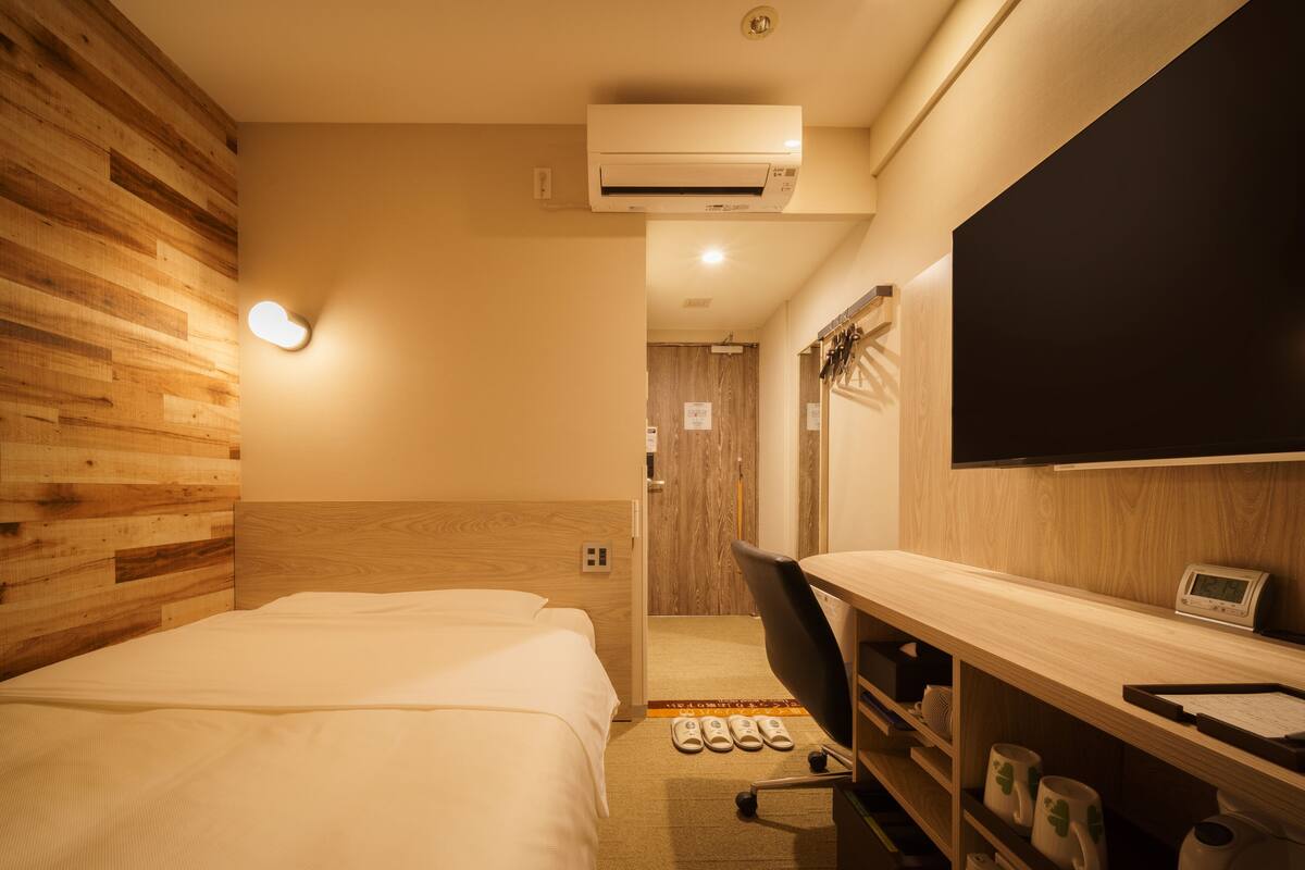 A hotel room with a wooden headboard, a bed, a desk with a chair, a TV, and a shoe rack.