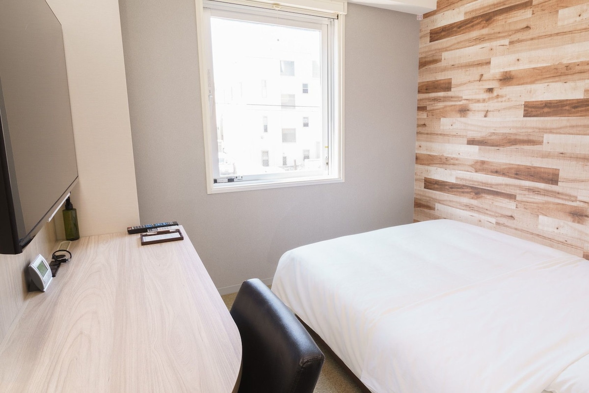 A hotel room with a bed, a desk with a chair, a window, and a wooden wall.