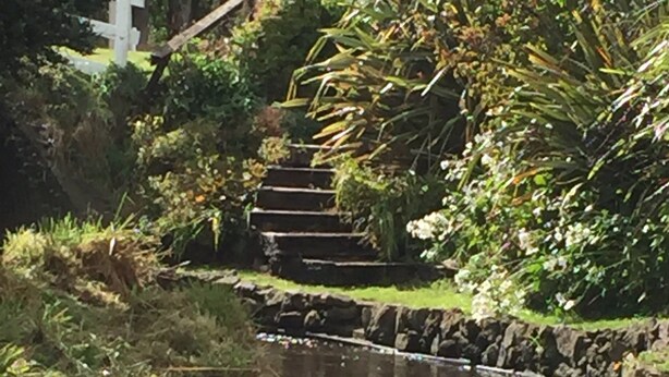 A stone staircase leading up to a white cross, surrounded by lush greenery and a small stream.