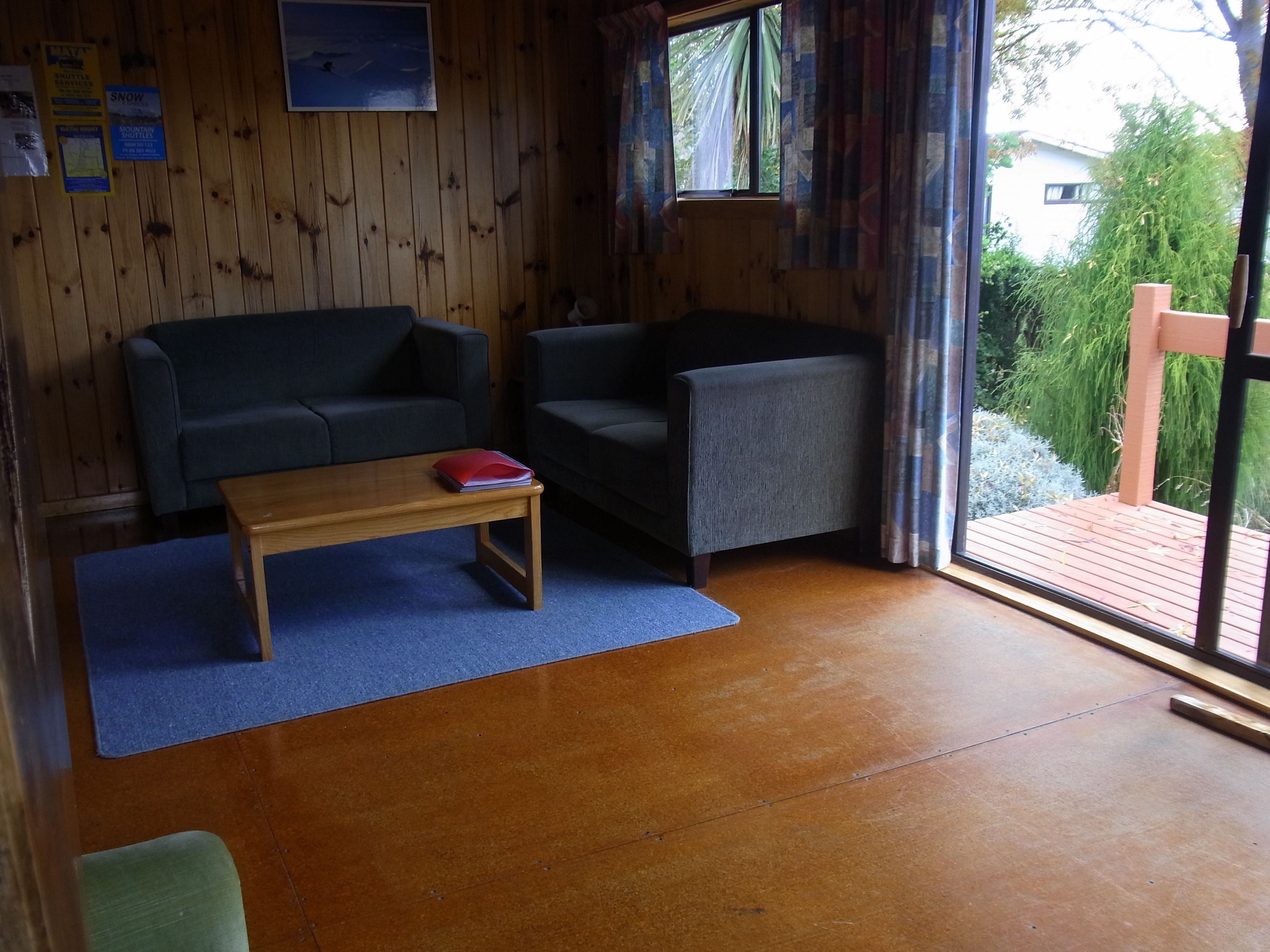 Una habitación con paredes de madera, alfombra azul, una mesa de centro de madera, dos sofás grises y una ventana con cortinas.