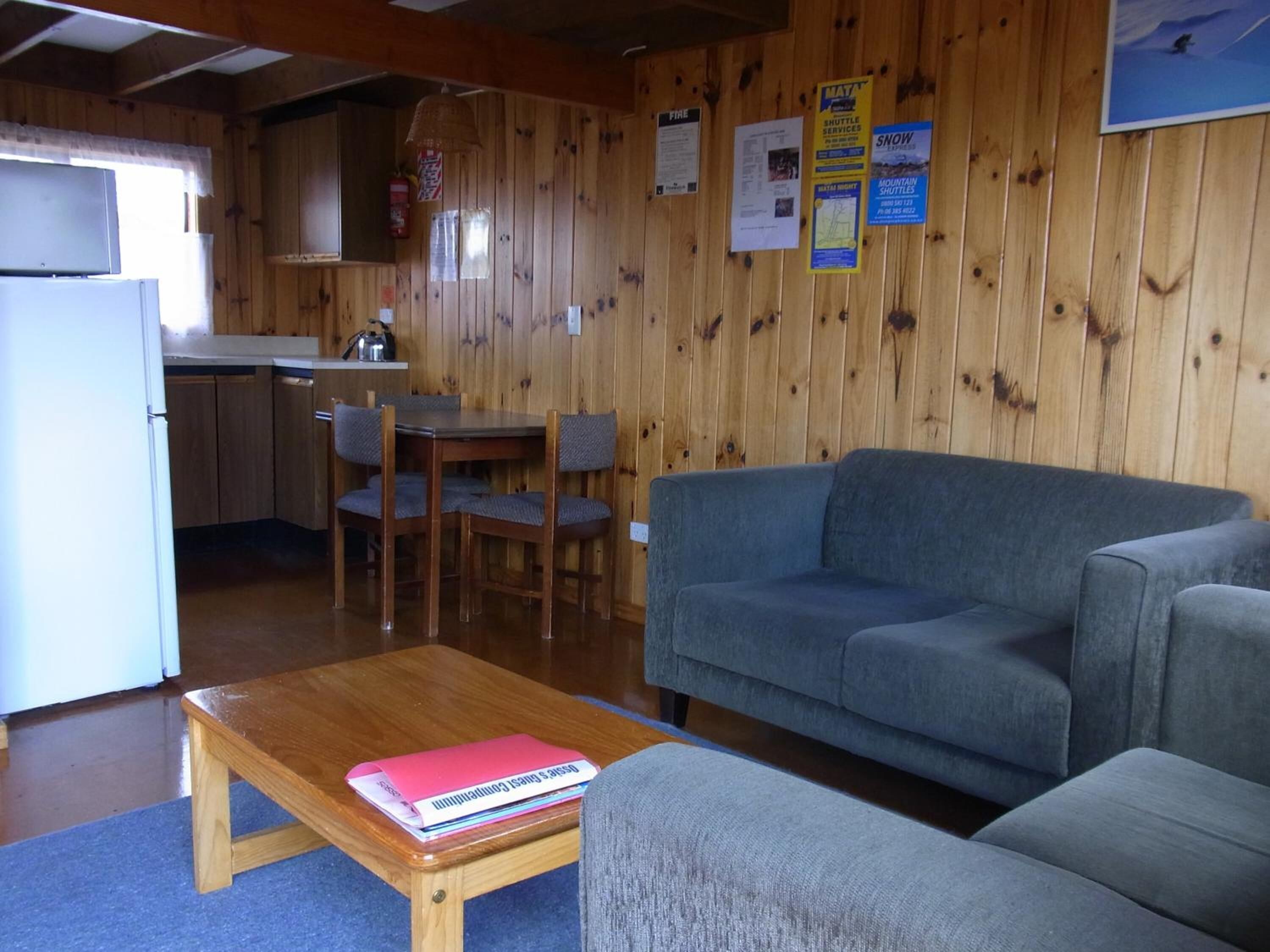 A room with a wooden interior, a grey sofa, a wooden table with a book, and a kitchen area with a refrigerator and a sink.