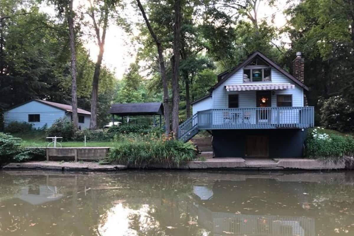 A lakeside cabin with a balcony, surrounded by trees and a calm water body.