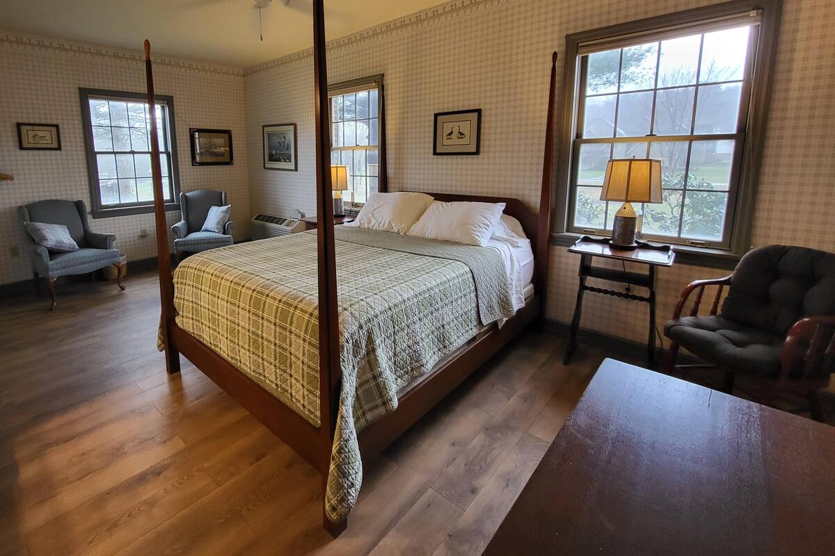 A four-poster bed with a quilt, two armchairs, a table with a lamp, and a view of the outdoors through a large window.