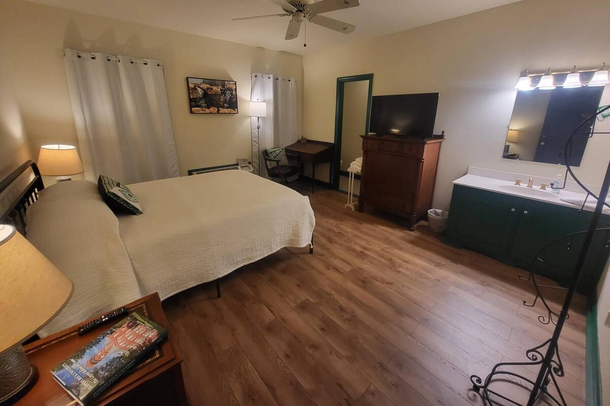 A hotel room with a bed, desk, chair, vanity with mirror, and a ceiling fan.