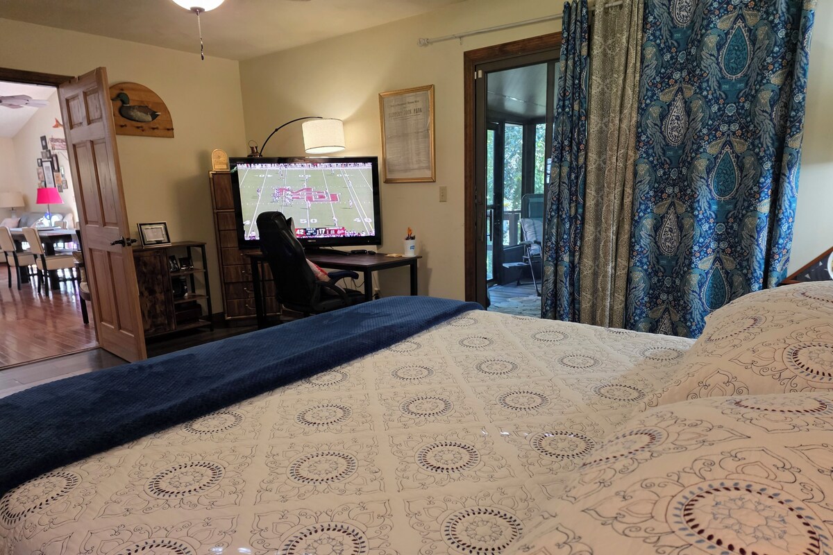 Una habitación con una cama, un escritorio con computadora, una silla y una puerta que da a otra habitación.