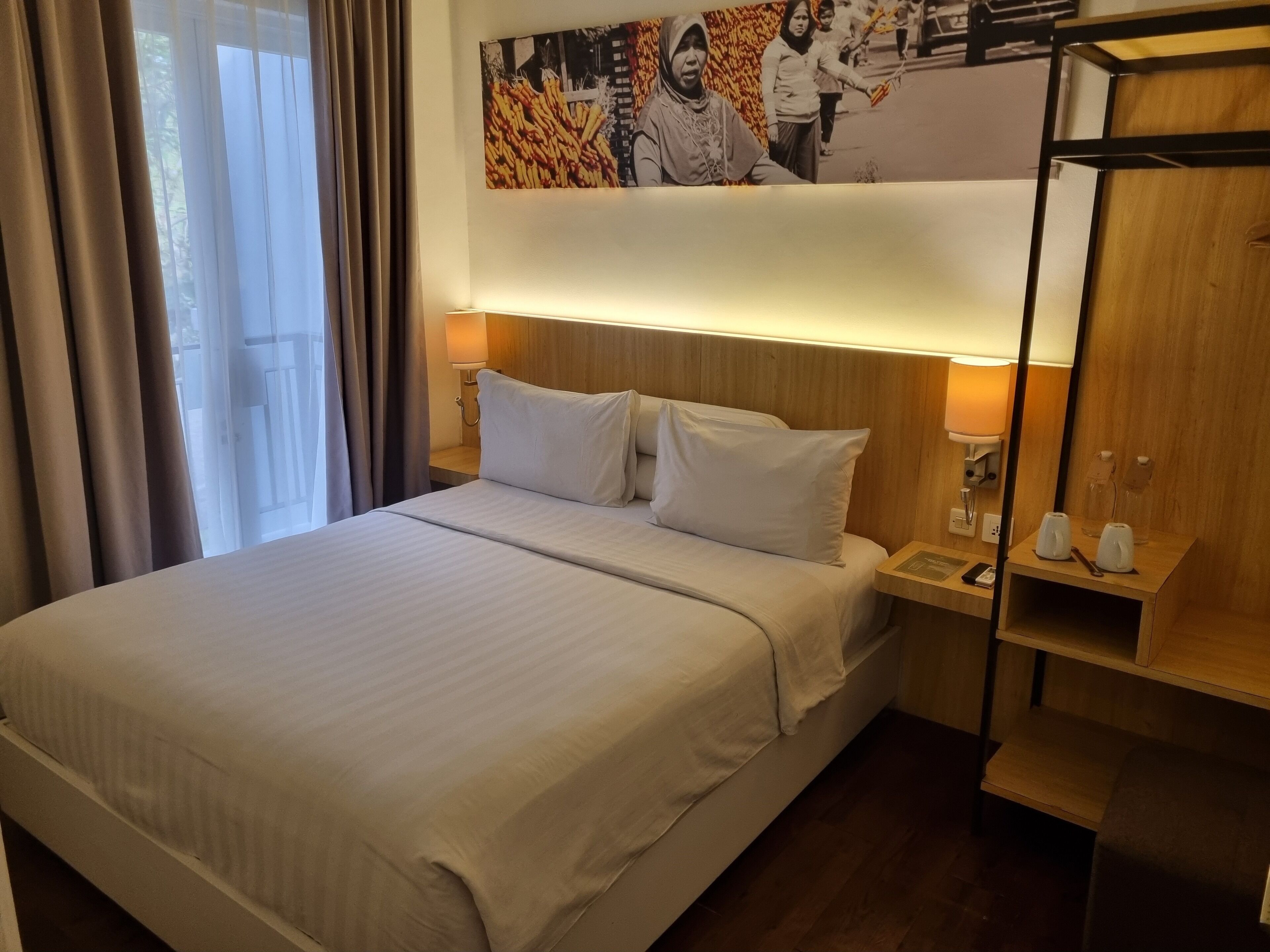 Ein Hotelzimmer mit einem großen Bett, einem Nachttisch mit einer Lampe, einem Holzregal mit einer Kaffeemaschine und einem an der Wand hängenden Bild.