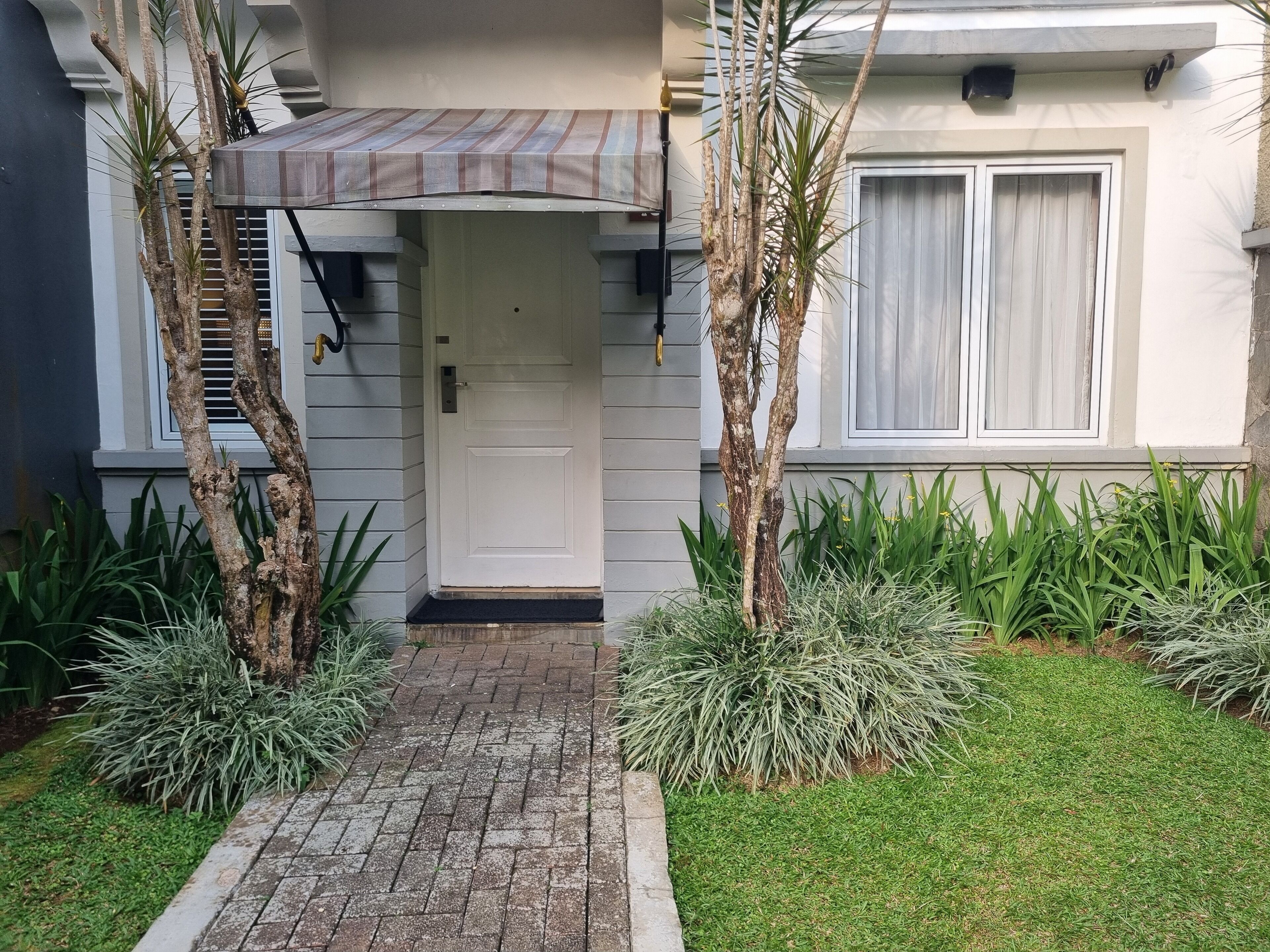 A house entrance with a white door, a small porch with a striped awning, and a brick pathway leading to the door.
