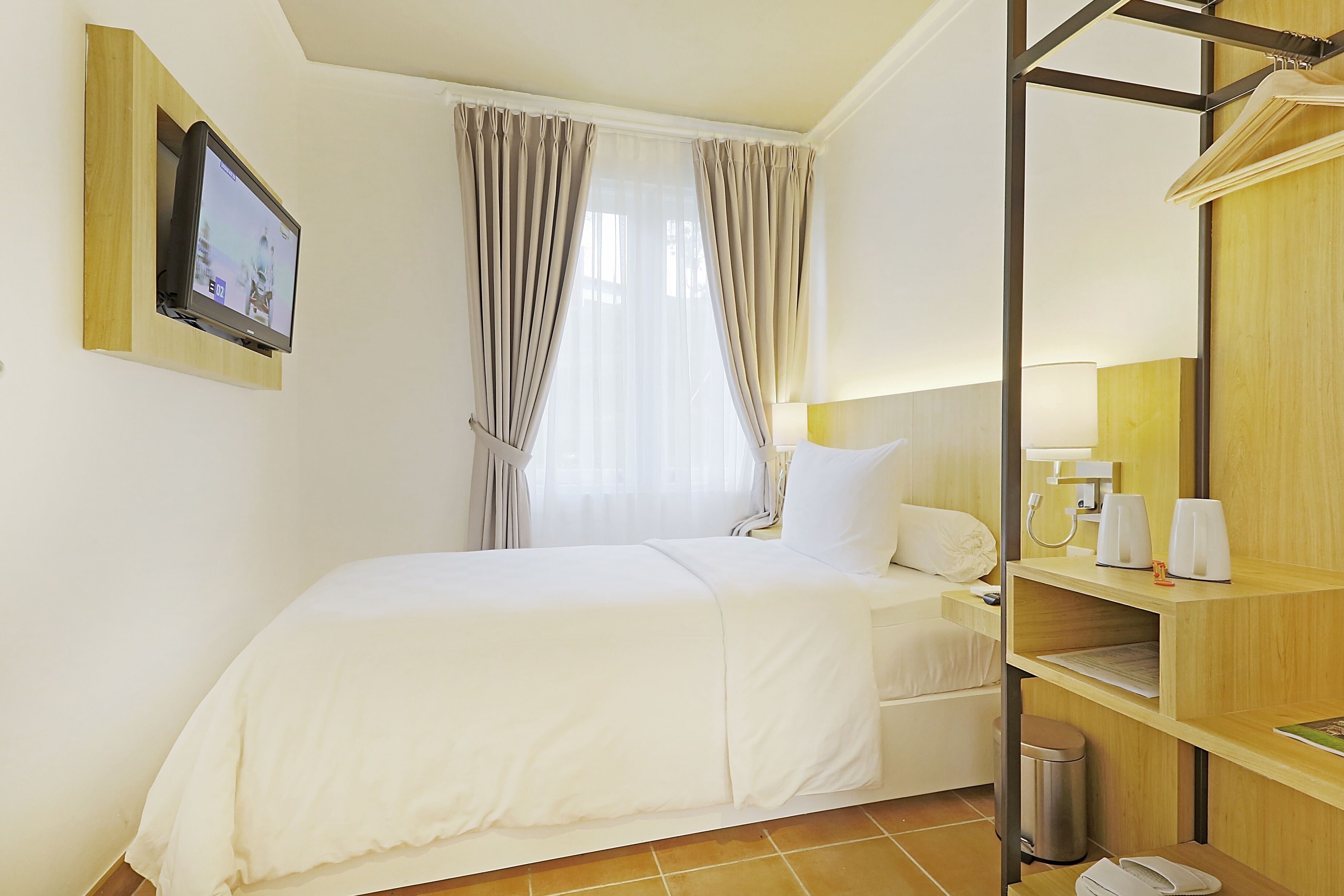 Habitación de hotel con cama, televisor montado en la pared, ventana con cortinas, mesita de noche con lámpara y estante con artículos de aseo.