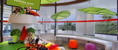 A modern hotel lobby with a large screen displaying the hotel name, plush toys on a tiered shelf, a colorful play area, and large windows offering a view of the outdoors.
