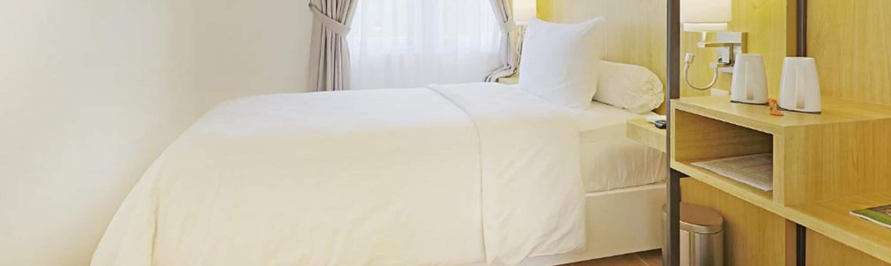 A hotel room with a bed, a nightstand, a trash bin, and a wooden shelf with a lamp and a cup.