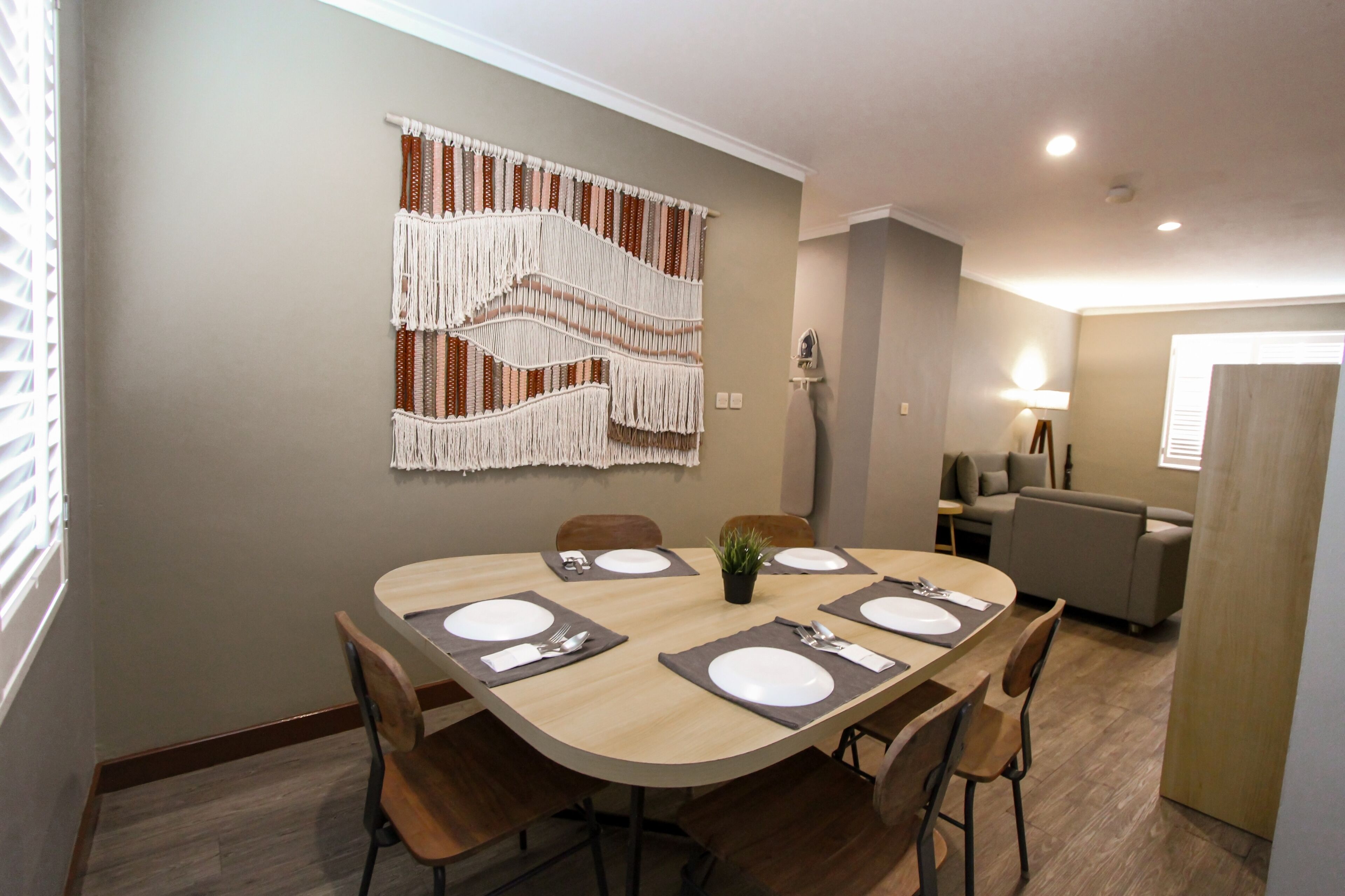 Un coin repas avec une table ronde dressée pour quatre personnes, entourée de chaises en bois. Une décoration murale en macramé est accrochée au mur, au-dessus de la table.
