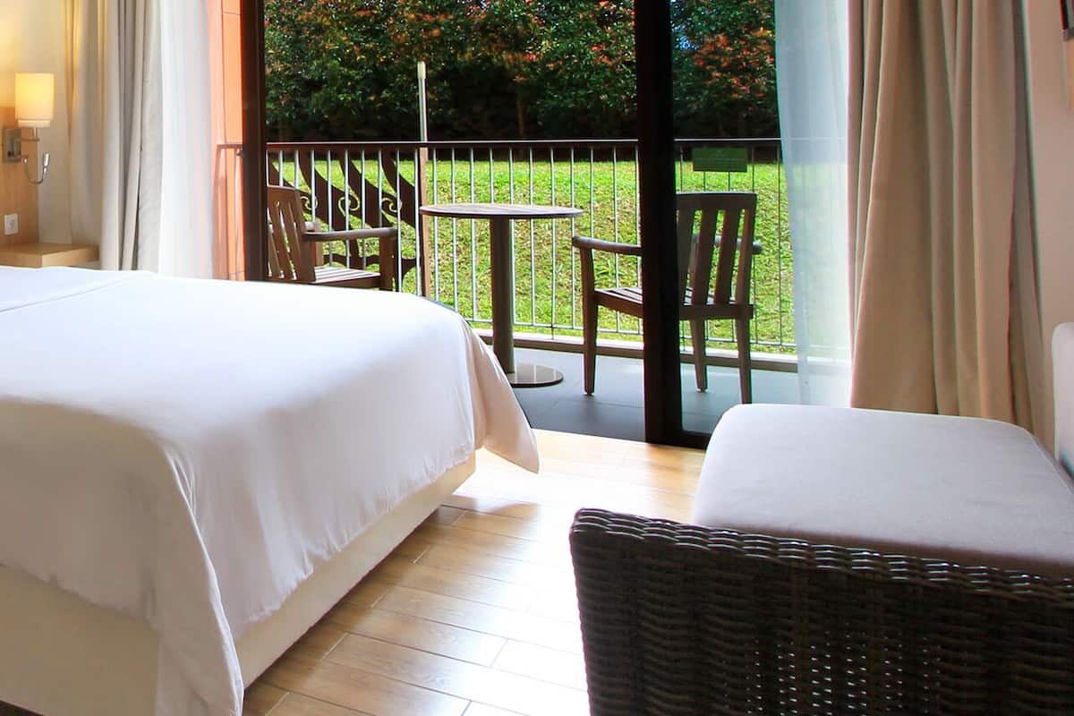 Quarto de hotel com uma cama, uma mesa com garrafa de água e cafeteira, uma cadeira de vime e uma varanda com vista para o verde.