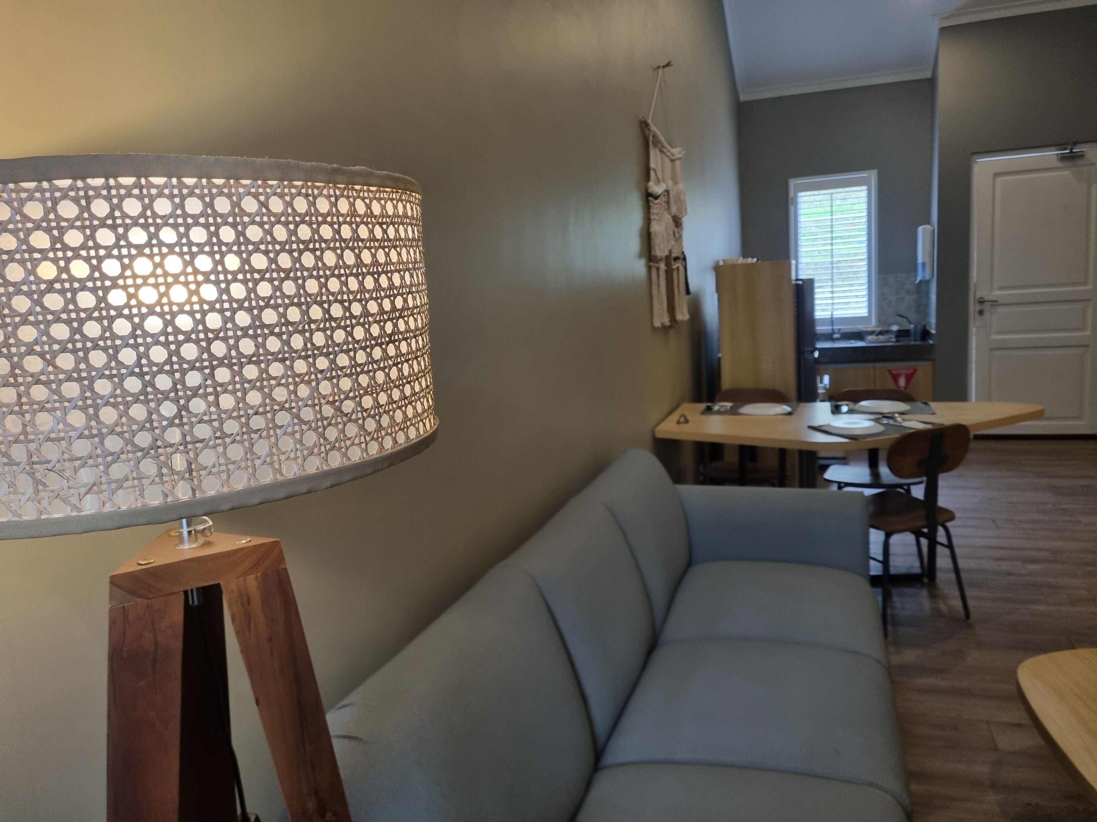 Una sala moderna con un sofá gris, una lámpara de pie de madera, una mesa de comedor con sillas y una cocina al fondo.