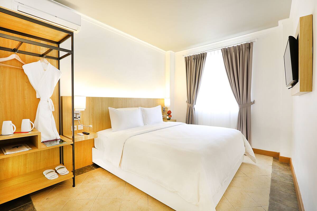 Quarto de hotel com uma cama bem arrumada, um guarda-roupa de madeira com roupão e chinelos, uma mesa de cabeceira com luminária e uma TV fixada na parede.