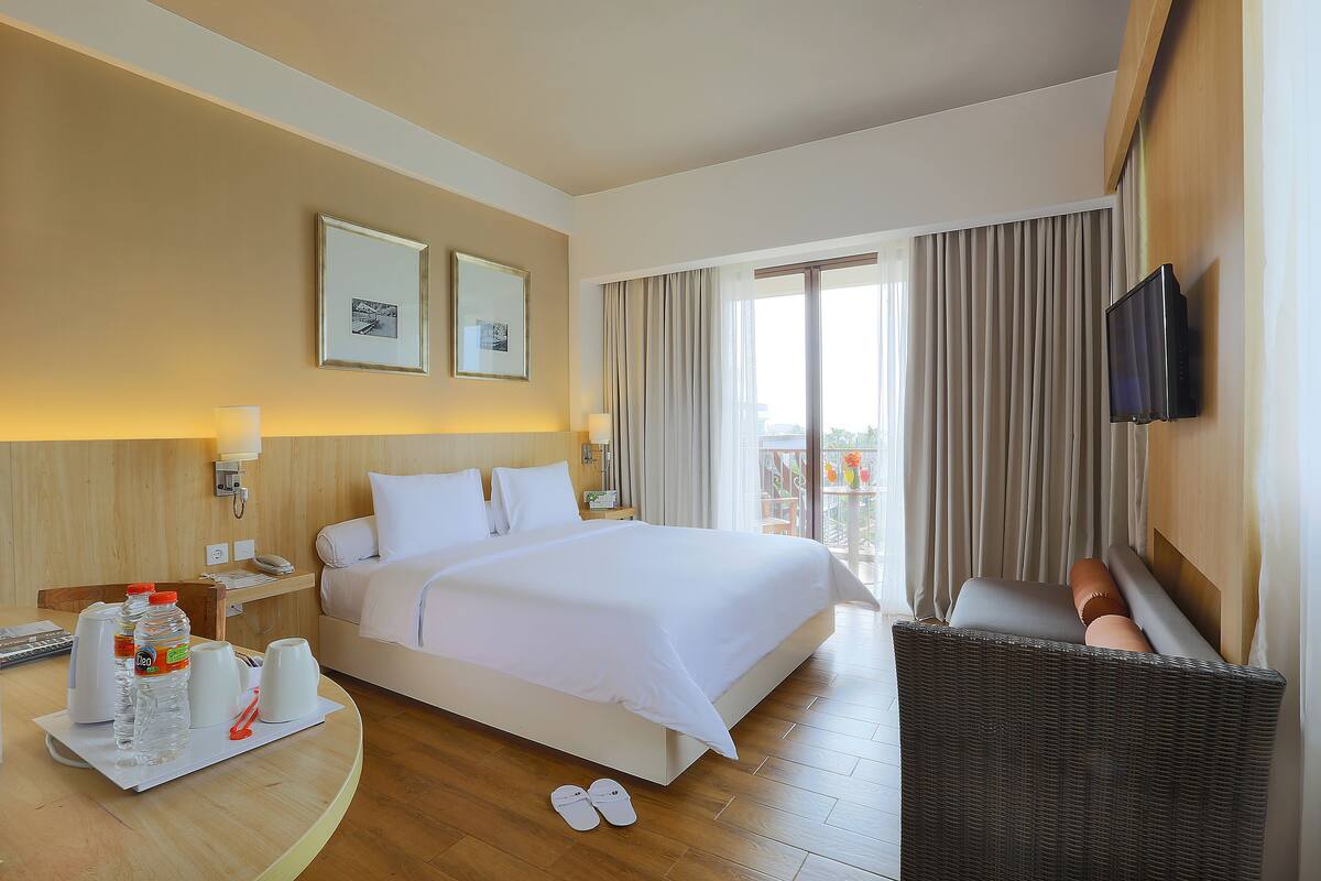 Quarto de hotel com cama, televisão na parede, varanda com cortinas e cadeira de vime.
