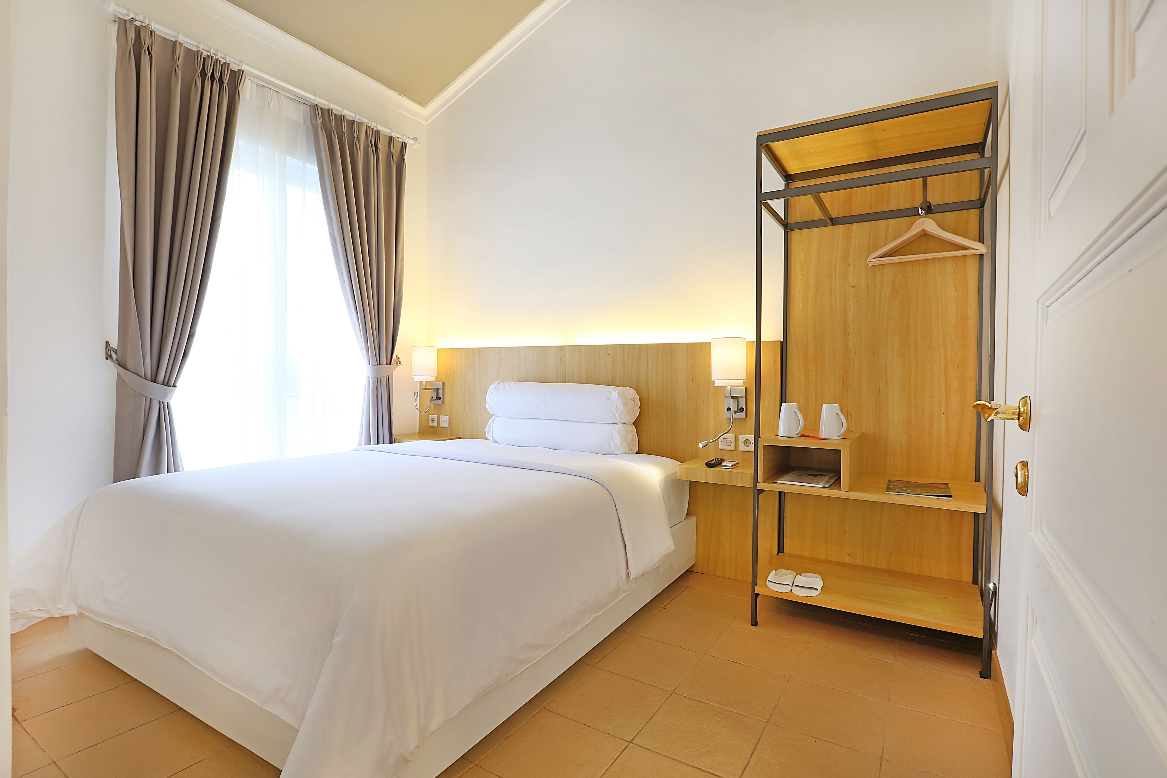 A hotel room with a bed, bedside lamps, a wooden wardrobe, and a shoe rack.